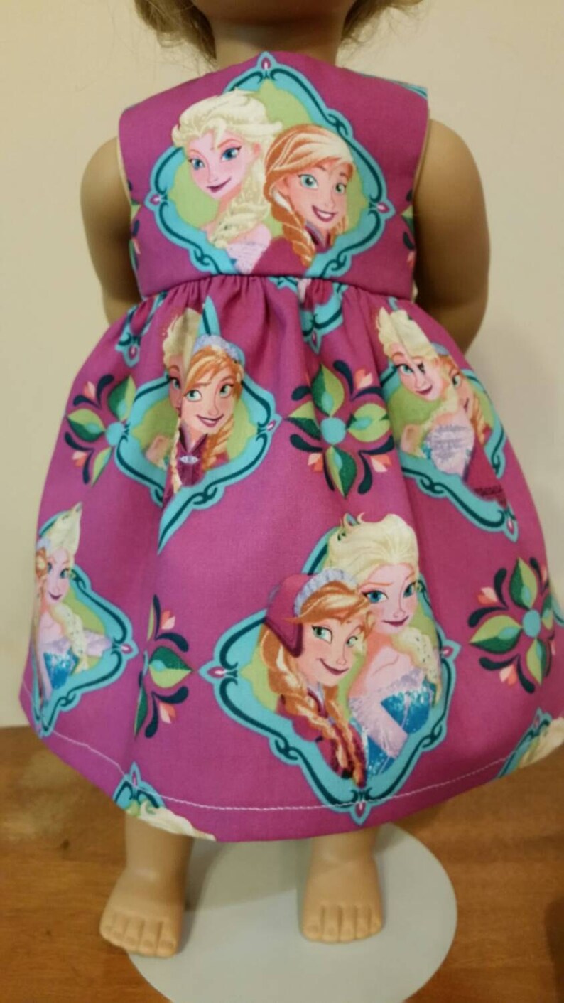May include: A sleeveless magenta dress featuring a repeating pattern of Elsa and Anna from Frozen. The dress has a scoop neckline and a gathered skirt. The characters are framed in teal and green decorative borders.