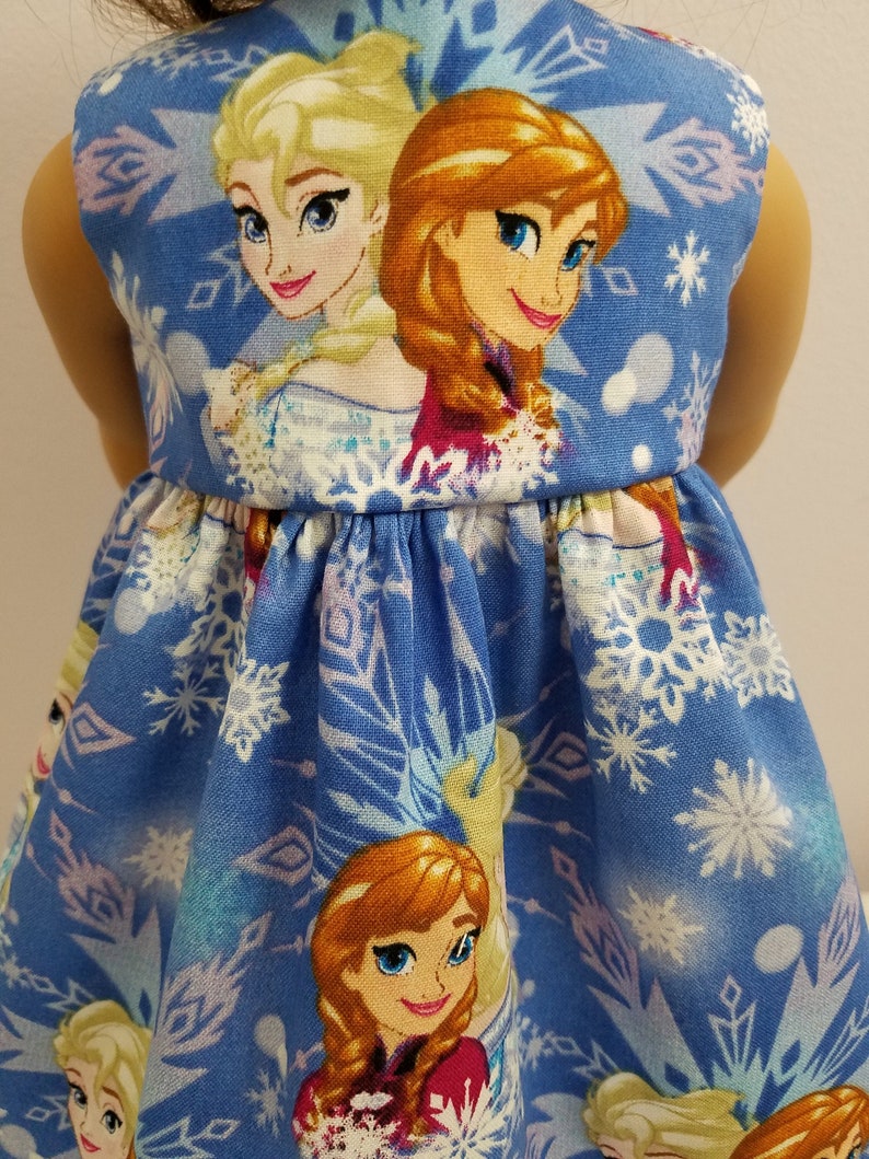 May include: A blue doll dress featuring the characters Elsa and Anna from the movie Frozen. The dress has a sleeveless design with a gathered waist and is decorated with snowflakes and images of the characters.
