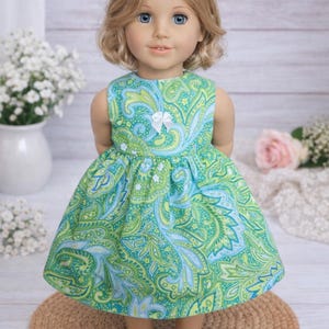 May include: A doll wearing a sleeveless green and blue paisley print dress with a white bow. The doll has blonde hair and blue eyes. The dress is knee-length and has a full skirt. The doll is standing on a round, woven mat.