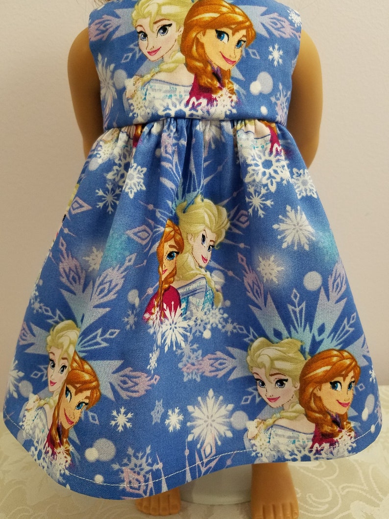May include: A blue doll dress featuring the characters Elsa and Anna from the movie Frozen, with a snowflake pattern. The sleeveless dress has a gathered waist and is made of fabric.