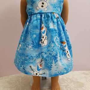 May include: A blue doll's dress with a pattern of Olaf the snowman from the film Frozen. The dress has a gathered skirt and a sleeveless design. The fabric background features a winter forest scene.