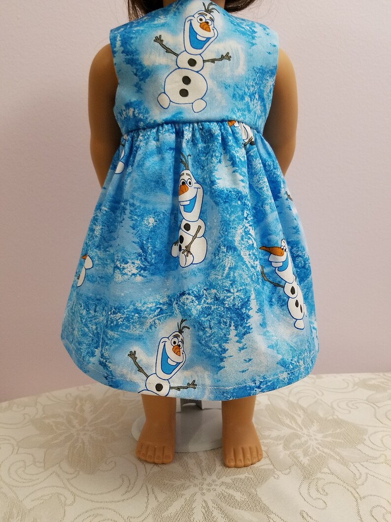 May include: A blue doll's dress with a pattern of Olaf the snowman from the film Frozen. The dress has a gathered skirt and a sleeveless design. The fabric background features a winter forest scene.