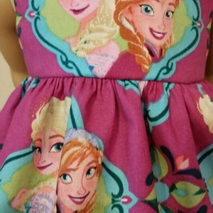 May include: A magenta dress featuring a repeating pattern of Elsa and Anna from the movie Frozen. The characters are framed in teal and white decorative borders. The dress has a gathered waist and a sleeveless design.