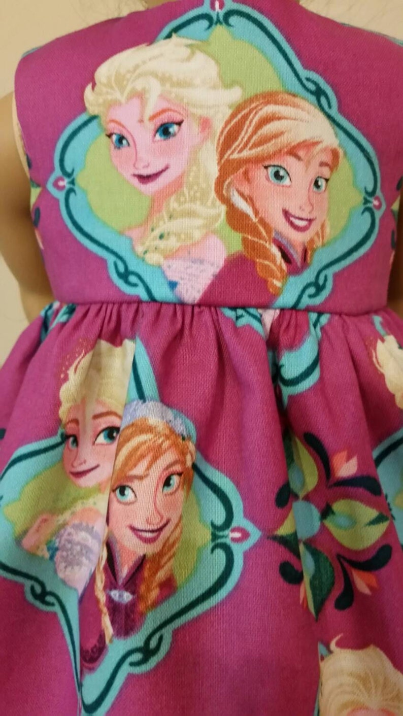 May include: A magenta dress featuring a repeating pattern of Elsa and Anna from the movie Frozen. The characters are framed in teal and white decorative borders. The dress has a gathered waist and a sleeveless design.