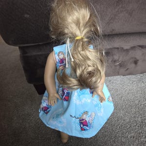 May include: A doll wearing a light blue dress featuring images of the characters Anna and Elsa from the movie Frozen. The dress has a snowflake pattern and is tied at the shoulders with white string. The doll has blonde hair tied back with a yellow band.