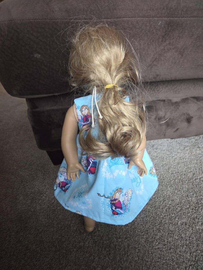 May include: A doll wearing a light blue dress featuring images of the characters Anna and Elsa from the movie Frozen. The dress has a snowflake pattern and is tied at the shoulders with white string. The doll has blonde hair tied back with a yellow band.