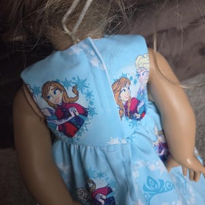 May include: A light blue doll dress featuring Anna and Elsa from the movie Frozen, with snowflake and character prints. The dress has a gathered skirt and is designed for a doll.
