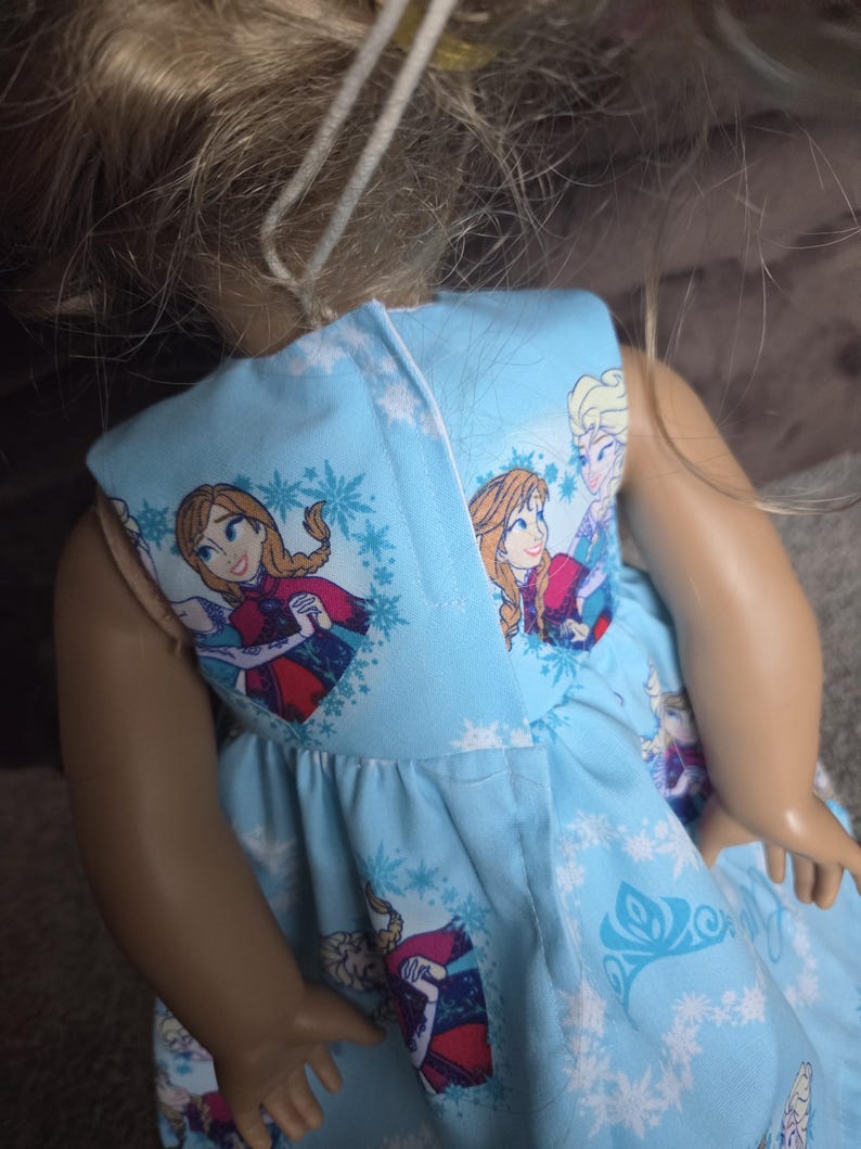 May include: A light blue doll dress featuring Anna and Elsa from the movie Frozen, with snowflake and character prints. The dress has a gathered skirt and is designed for a doll.