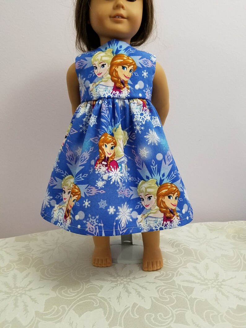 May include: A blue doll dress featuring images of animated characters and snowflakes. The sleeveless dress has a flared skirt and is designed for a doll. The fabric is a vibrant blue with a repeating pattern of the characters and snowflakes.