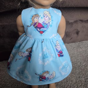 May include: A light blue doll dress featuring a heart-shaped graphic with two cartoon characters. The dress has a flared skirt with repeating character and snowflake patterns. The doll is standing on a gray surface.