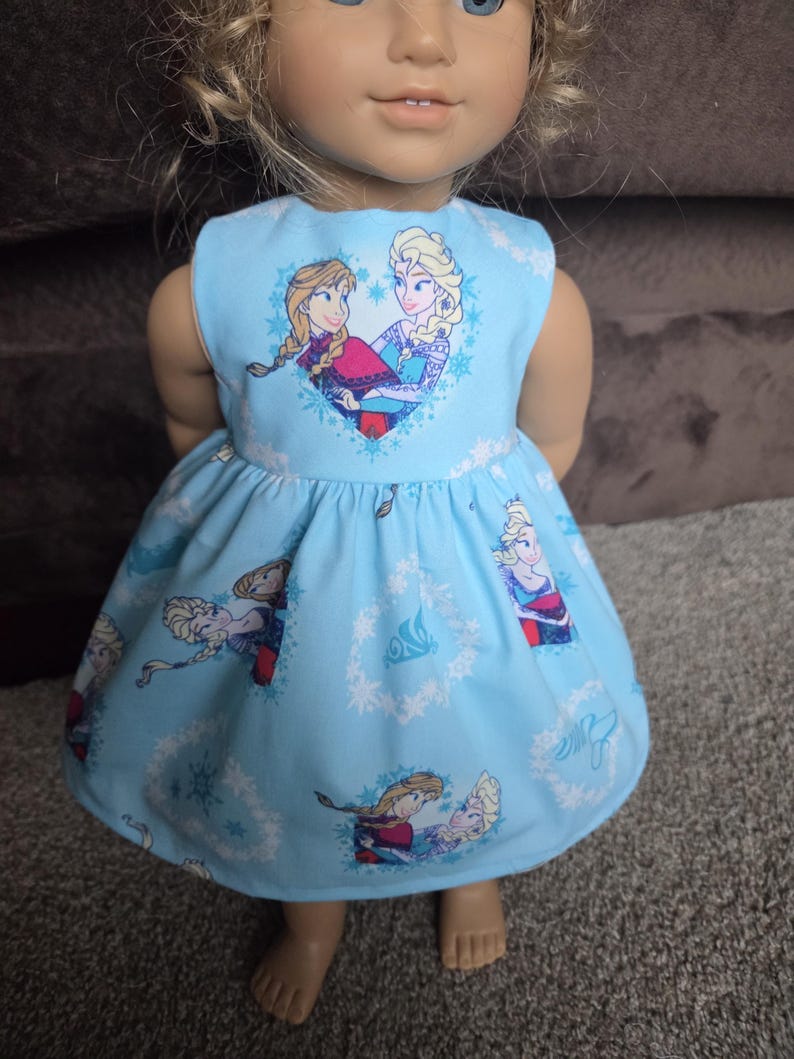 May include: A light blue doll dress featuring a heart-shaped graphic with two cartoon characters. The dress has a flared skirt with repeating character and snowflake patterns. The doll is standing on a gray surface.