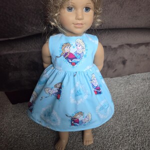 May include: A doll wearing a light blue dress featuring a print of two cartoon characters. The sleeveless dress has a heart-shaped design on the bodice and a flared skirt. The doll has blonde hair and blue eyes.