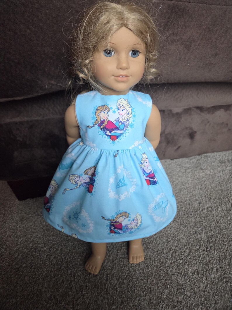 May include: A doll wearing a light blue dress featuring a print of two cartoon characters. The sleeveless dress has a heart-shaped design on the bodice and a flared skirt. The doll has blonde hair and blue eyes.