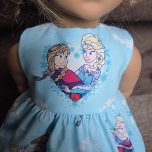 May include: A light blue doll dress featuring a print of two animated characters in a heart-shaped design. The dress has a sleeveless design and a flared skirt, with additional character and snowflake patterns.