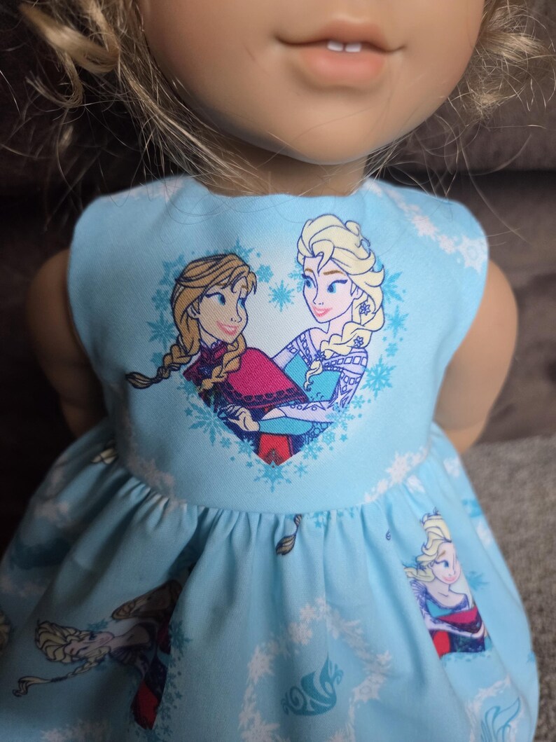 May include: A light blue doll dress featuring a print of two animated characters in a heart-shaped design. The dress has a sleeveless design and a flared skirt, with additional character and snowflake patterns.