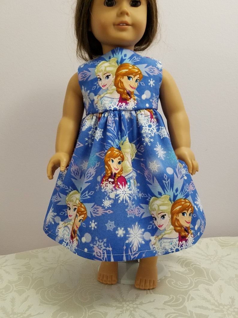 May include: A blue doll dress featuring images of two animated characters, with a snowflake pattern. The sleeveless dress has a fitted bodice and a flared skirt. The characters are blonde and auburn-haired women.