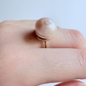 Peut inclure: Une bague dorée ornée d'une grande sphère ronde blanc rosé nacré, surmontée d'un petit accent sphérique blanc distinct. L'anneau est fin et métallique, soutenant la structure centrale composée de deux éléments arrondis superposés.