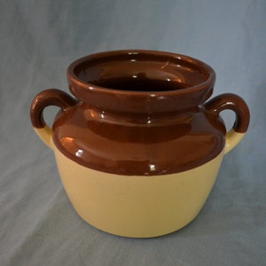 Op de afbeelding: Een tweekleurige keramische pot met een bruine bovenkant en handvatten en een crèmekleurige basis. De pot heeft een brede opening en twee gebogen handvatten. De glanzende afwerking reflecteert licht en benadrukt het gladde oppervlak. Deze decoratieve pot is geschikt voor keuken of woondecoratie.