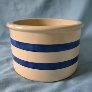 May include: A cream-colored ceramic crock with two horizontal blue stripes. The open-top container has a rounded shape and a slightly flared rim. The stripes are evenly spaced around the body of the crock.