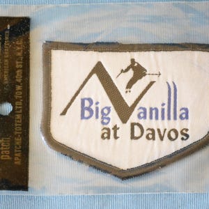 May include: A vintage ski patch in a clear plastic sleeve. The patch is shield-shaped with a white background and a brown border. It features a skier graphic and the text "Big Vanilla at Davos."