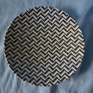 May include: Round ceramic plate with a cream-colored base and a repeating navy blue zigzag pattern. The plate is centered on a light blue fabric background, creating a simple, clean aesthetic.