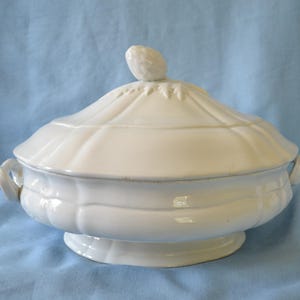 May include: A white ceramic soup tureen with a lid and handles. The tureen has a fluted design and a small knob on top of the lid. The tureen is on a light blue background.