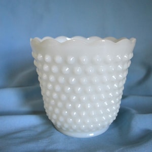 May include: A white hobnail glass vase with a scalloped rim. The vase has a cylindrical shape and is covered in small, raised, rounded bumps. The background is a solid light blue fabric.