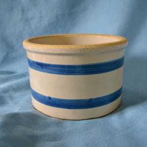 May include: A cylindrical ceramic crock with a cream-colored base and two horizontal blue stripes. The rim is a light yellow color. The crock is likely used for food storage or decoration.