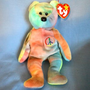 May include: A colorful tie-dye teddy bear plush toy with a peace sign on its chest. The bear has a red heart-shaped tag with a yellow star. The bear's colors include orange, yellow, green, and blue.