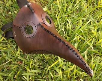 May include: A brown leather plague doctor mask with a long beak and two round eyeholes. The mask has a stitched design and is lying on a bed of green grass.