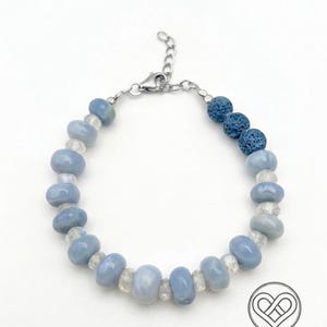 May include: A beaded bracelet featuring smooth, round, light blue beads alternating with small, clear, faceted spacer beads. Three dark blue, porous, textured lava stone beads are positioned on one side. The bracelet has a silver-toned metal clasp.