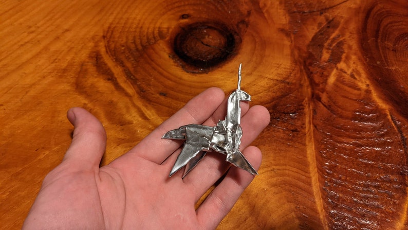 May include: A silver origami unicorn, folded from a single sheet of aluminium foil, is held in a person's hand. The unicorn is standing on its four legs, with its horn pointing upwards.