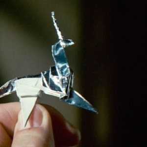 May include: A silver origami unicorn with a pointed horn. The unicorn is folded from a single sheet of paper and is held in a person's hand.