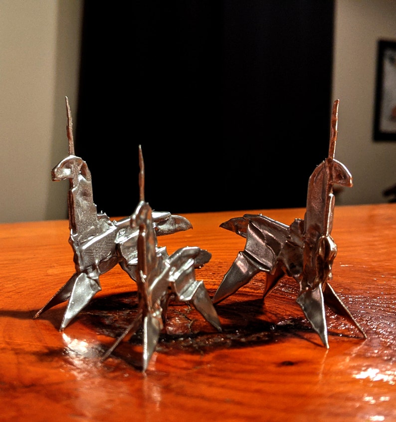 May include: Three silver origami unicorns stand on a wooden surface. The unicorns are made of folded metal and have a shiny finish.