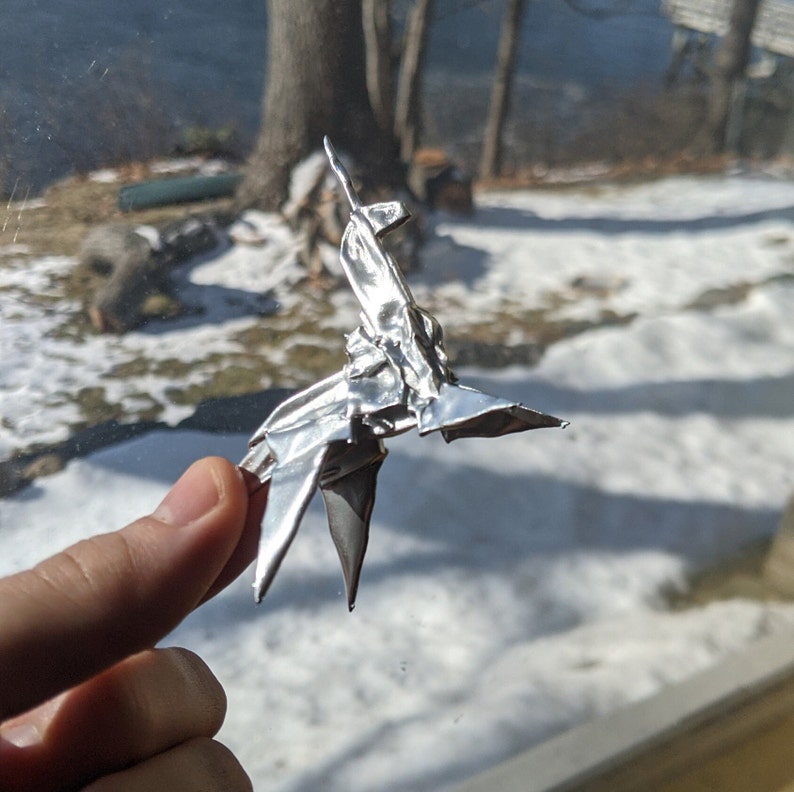 May include: A silver origami unicorn, folded from aluminium foil, is held in a person's hand. The unicorn is standing on its hind legs with its horn pointing upwards.
