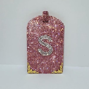 May include: A pink pendant encrusted with sparkling crystals. The pendant features a large letter "S" in the center, outlined with smaller, clear crystals. Gold-colored corner accents add a touch of elegance. The bail is also covered in pink crystals.