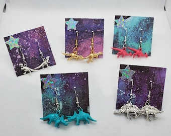 Könnte beinhalten: Sechs Paare Dinosaurier-Ohrringe, die auf Karten mit Galaxie-Druck präsentiert werden. Die Ohrringe sind silber, gold, pink und blau.