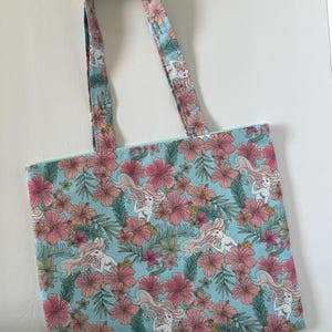 Peut inclure: Un sac fourre-tout bleu clair avec un motif r&eacute;p&eacute;t&eacute; de fleurs d'hibiscus roses, de feuilles vertes et de sir&egrave;nes. Les poign&eacute;es reprennent le motif. Le sac en tissu est con&ccedil;u pour transporter des objets.