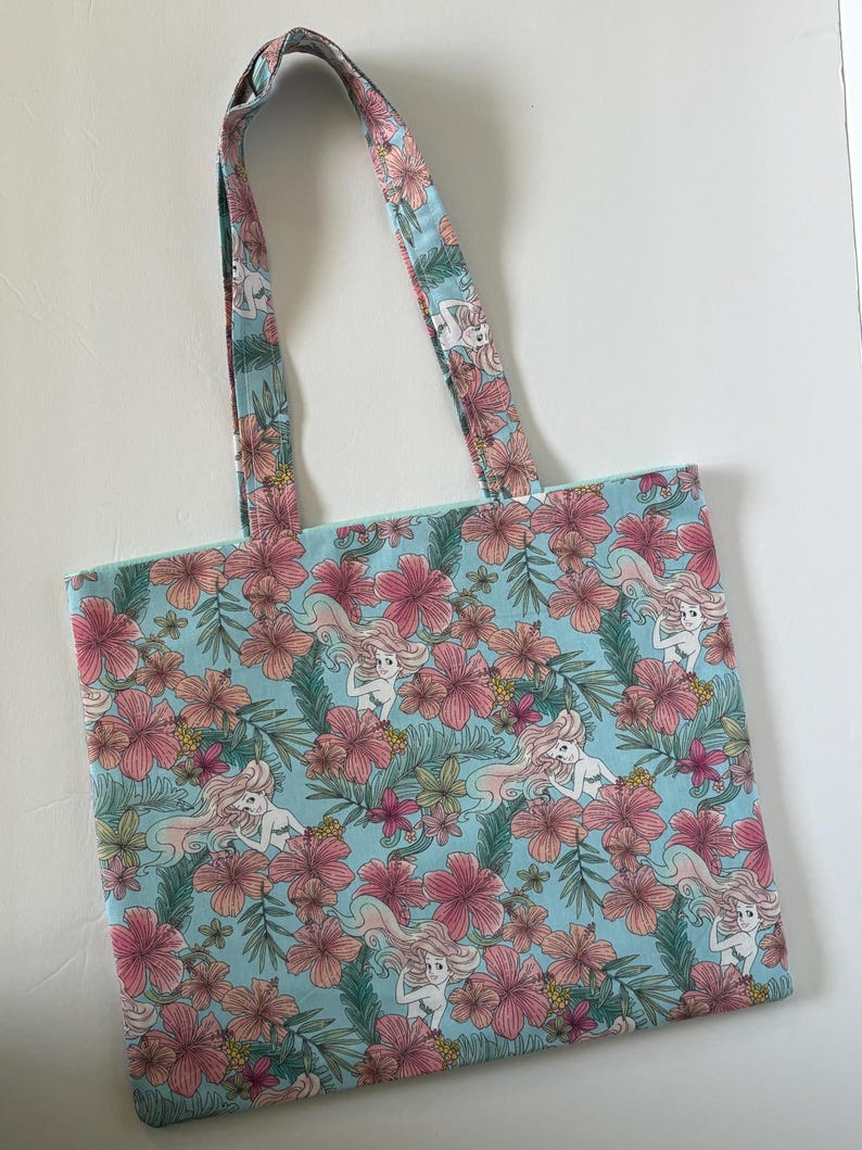 Peut inclure: Un sac fourre-tout bleu clair avec un motif r&eacute;p&eacute;t&eacute; de fleurs d'hibiscus roses, de feuilles vertes et de sir&egrave;nes. Les poign&eacute;es reprennent le motif. Le sac en tissu est con&ccedil;u pour transporter des objets.