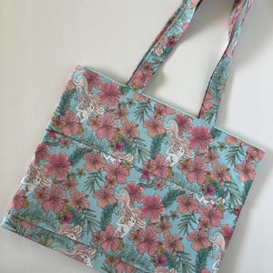 Peut inclure: Un sac fourre-tout bleu clair avec un motif floral et sir&egrave;ne. Le sac pr&eacute;sente des fleurs d'hibiscus roses, des feuilles vertes et des illustrations de sir&egrave;nes. Les poign&eacute;es ont le m&ecirc;me motif que le sac. Le sac est en tissu.