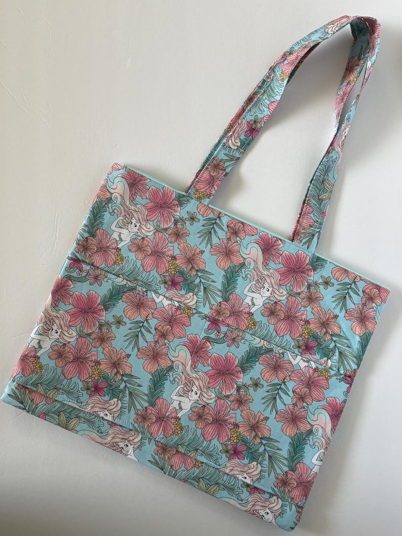 Peut inclure: Un sac fourre-tout bleu clair avec un motif floral et sir&egrave;ne. Le sac pr&eacute;sente des fleurs d'hibiscus roses, des feuilles vertes et des illustrations de sir&egrave;nes. Les poign&eacute;es ont le m&ecirc;me motif que le sac. Le sac est en tissu.