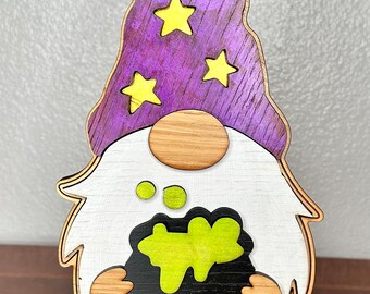 May include: A wooden gnome with a purple hat and yellow stars, holding a black cauldron with green liquid inside. The gnome is standing on a black base.