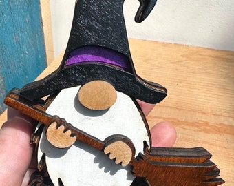May include: A wooden witch gnome ornament with a black hat, purple trim, white beard, and brown broom.