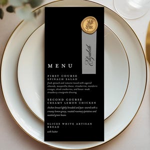 May include: A formal place setting with a black menu card featuring gold accents. The menu lists a spinach salad and creamy lemon chicken. Gold flatware and white plates with gold rims complete the elegant table setting.