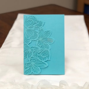 May include: A turquoise blue card with a delicate floral design cut out of the top right corner. The card is on a white ruffled surface.