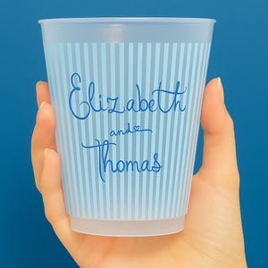 Op de afbeelding: Een doorzichtige plastic beker met lichtblauwe verticale strepen en de namen "Elizabeth and Thomas" in een sierlijk lettertype. De beker wordt in een hand gehouden tegen een blauwe achtergrond.