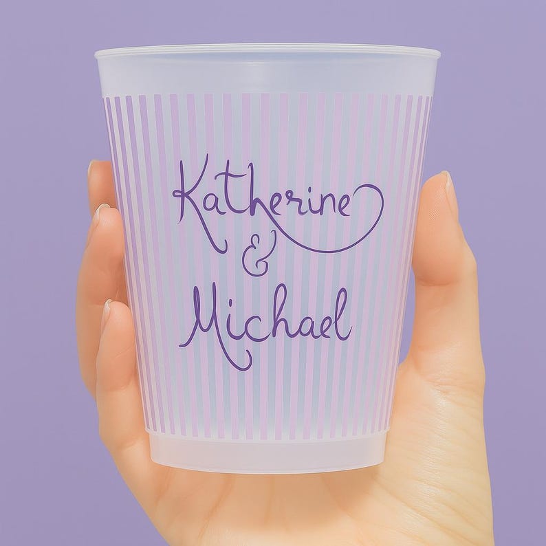 Op de afbeelding: Een doorzichtige plastic beker met verticale strepen in lavendel en lichtroze. De beker is gepersonaliseerd met de namen "Katherine & Michael" in een cursief lettertype. De beker wordt door een hand tegen een lavendelkleurige achtergrond gehouden.