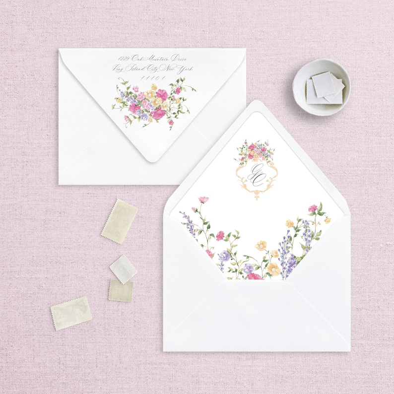 May include: Two white envelopes with floral designs. The envelope on the left has a floral design with pink, yellow, and purple flowers. The envelope on the right has a floral design with pink, yellow, and purple flowers and a monogram in the center. The address on the left envelope reads "629 Oak Mountain Drive, Bay Island City, New York 11101."