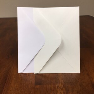 May include: Two white envelopes, one slightly larger than the other, are stacked on a wooden surface.