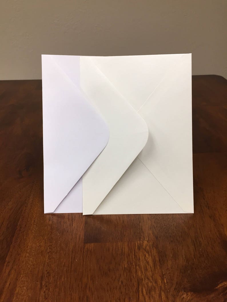 May include: Two white envelopes, one slightly larger than the other, are stacked on a wooden surface.
